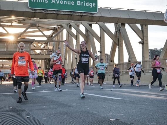 Runners take part in a road race on a bridge beneath a Willis Avenue Bridge sign. The sun is shining, and one runner in the centre is smiling and waving at the camera. Other runners are seen in various athletic outfits.